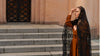 Woman in a brown dress and black headscarf standing on steps with a beige wall in the background